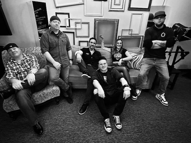 Black and white photo of the six members of The Benjamins posing around a couch backstage